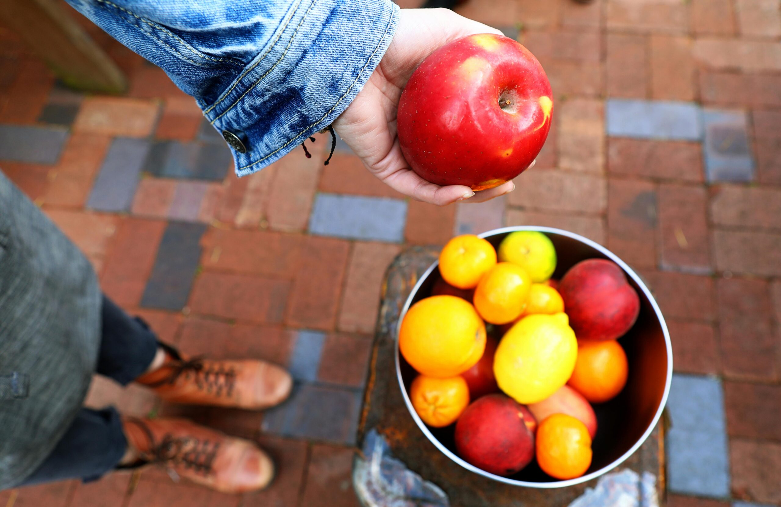 Nosotros Top view of a person holding a red apple above a bowl of fresh fruits on a brick surface.