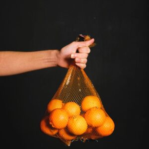 A hand holding a mesh bag filled with vibrant fresh oranges against a dark background.