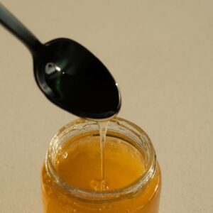A close-up of golden honey dripping from a black spoon into a jar, captured in bright lighting.